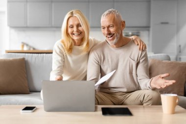 Mutlu yetişkin erkek ve kadın internet faturalarını ödüyor, evdeki koltukta oturuyor, dizüstü bilgisayar kullanıyor, evrakları tutuyor, mektuplar yazıyor, sanal bankacılıktan hoşlanan yaşlı çift.