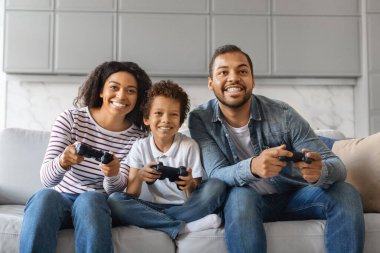 Neşeli siyahi aile, ebeveynler ve küçük oğullar evdeki video oyunlarında birbirleriyle rekabet ediyorlar, pozitif Afrikalı anne, baba ve erkek çocuk joystickler kullanıyor, oturma odasında eğleniyorlar.