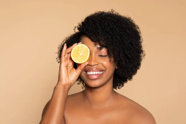 Radiant young black woman smiling with eyes closed, holding fresh lemon slice over her eye, symbolizing natural beauty and skincare, against beige backdrop