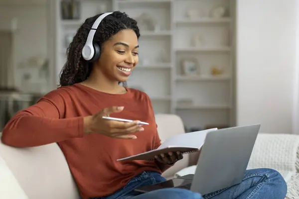 Video Call. Smiling Black Woman Wearing Headphones Teleconferencing On Laptop At Home, Happy African American Lady Talking And Gesturing At Computer Web Camera While Sitting On Couch, Free Space