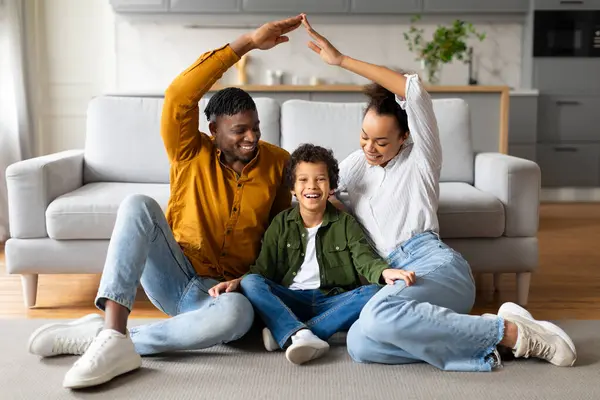 Happy black family sits on the floor making symbolic house shape with their hands, expressing unity and comfort in their bright and cozy living room