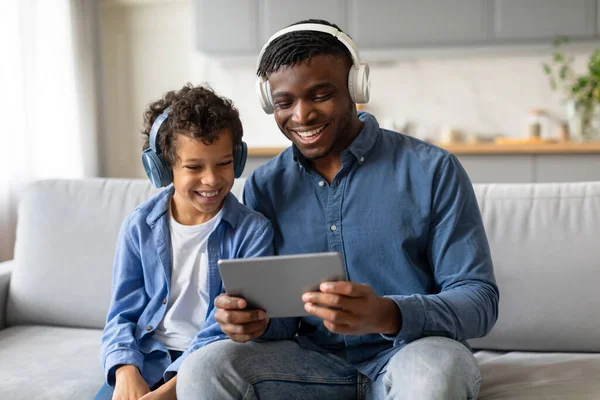 笑着带着耳机的黑人男孩和他快乐的父亲一起在平板电脑上享受着滑稽的视频 在舒适的沙发上分享着快乐的时光 — 图库照片