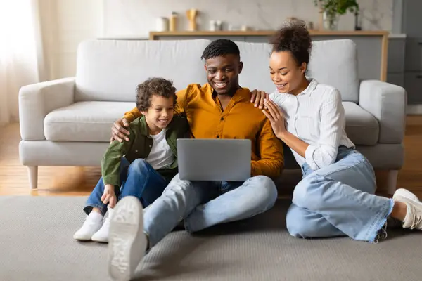 带着笔记本电脑坐在地板上笑着的黑人家庭 在家里一起参加在线活动 在电脑上上网 分享快乐时光 — 图库照片