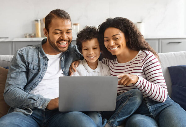 Happy black family with little son enjoying fun content on laptop while sitting comfortably on their couch in living room, cheerful african american parents and male kid using computer at home