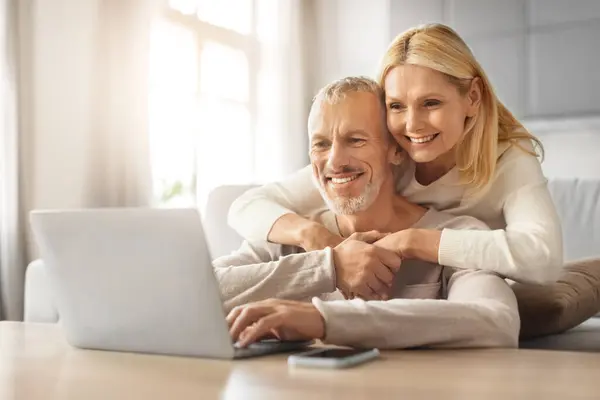 Loving european retired couple planning vacation together, using laptop computer at home, booking trip, hotel and flight online, embracing and smiling, copy space, sun flare