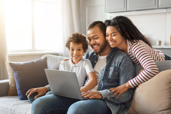 Happy young black family of three using laptop together at home, cheerful african american parents and preteen son browsing internet on computer or shopping online while relaxing on couch