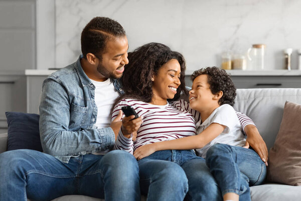 Relaxed black family lounging on the couch with remote in hand, happy young african american parents and male child enjoying watching TV together, having fun in cozy living room interior, closeup