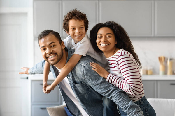 Портрет веселой чёрной семьи с маленьким сыном, обнимающим и улыбающимся перед камерой, счастливые афроамериканские родители, сближающиеся со своим сыном-подростком, позируя вместе в домашнем интерьере