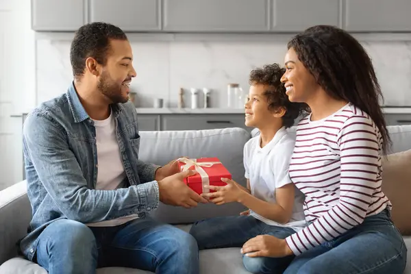 Portrait of little black son making surprise for his dad, giving present box, greeting happy man with Fathers day or birthday. African american family sitting on the couch in living room at home
