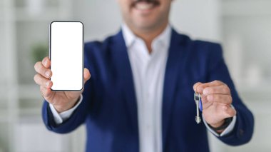 Mavi takım elbiseli gülümseyen bir Avrupalı işadamının elindeki boş ekran ile akıllı bir telefonun yakın plan görüntüsü. Arka planı bulanık ve ev anahtarları kesilmiş.