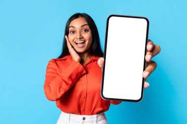 Wow başvuru, mobil teklif. Heyecanlı Hintli kadın, boş ekranlı büyük bir cep telefonu gösteriyor. Mavi stüdyo arkaplanının üzerinde duruyor. Boş dokunmatik ekranlı aleti tutarken yüze dokunuyor.