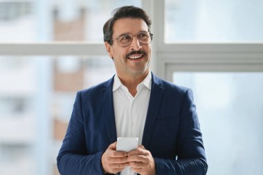 Beyaz gömlekli, elinde cep telefonu olan ve gülümseyen bir pencereden dışarı bakan iyimser, beyaz takım elbiseli beyaz bir işadamı.