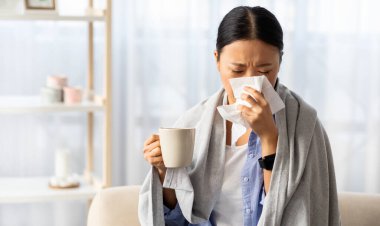 Hasta Asyalı kadın soğuk algınlığı ya da grip olmuş, sıcak battaniyeye sarılmış, hapşırıyor, sıcak içecek ve peçeteyle bardağı tutuyor, panoramada fotokopi çekiyor.