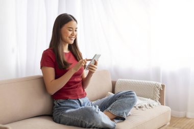 Güzel uygulama. Evde cep telefonu kullanan gülümseyen Asyalı kadın, neşeli güzel Koreli kadın sosyal medyayı kaydırıyor ya da akıllı telefondan alışveriş yapıyor, oturma odasında rahat koltukta oturuyor.