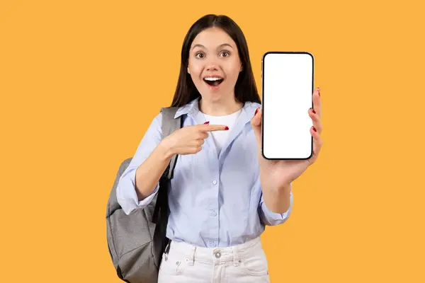 穿着蓝色衬衫的年轻女人感到惊讶 兴奋地指着空白的智能手机屏幕 背着黄色背景的背包 嘲讽着 — 图库照片