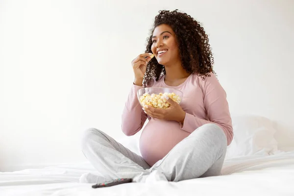 Happy black expectant mother in pink shirt enjoying bowl of popcorn while sitting cross-legged on white bed at home, smiling expectant mother watching movie or tv, enjoying domestic leisure