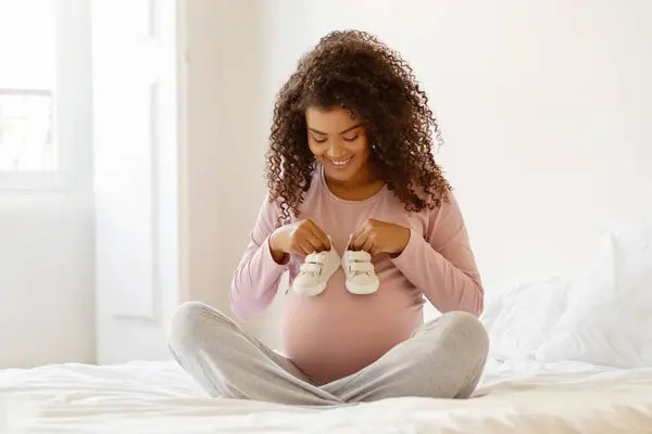 Mujer Embarazada Negro Sonriente Sentado Cama Sosteniendo Zapatillas Deporte Bebé —  Fotos de Stock