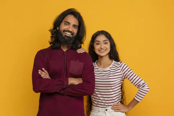 Positive young indian man and woman friends wearing casual clothing posing together on yellow studio background, smiling at camera. Teamwork, friendship, relationships concept