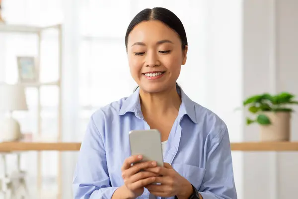 Portrait of happy attractive millennial chinese lady wearing blue shirt using cell phone at home, looking at smartphone screen and smiling, chatting with men on dating mobile app, copy space