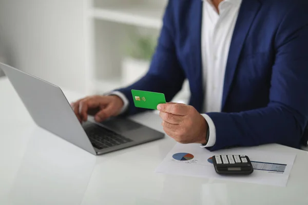 Mature caucasian businessman in a stylish blue blazer making an online payment with a green credit card while working on a laptop, with financial documents and a calculator on the desk