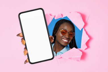 Yeni uygulama. Güneş gözlüklü neşeli Afro-Amerikan genç bayan akıllı telefonu boş ekranla sunuyor, elindeki aleti yırtılmış pembe kağıt arka planda gösteriyor. Mobil reklam. Kolaj, model