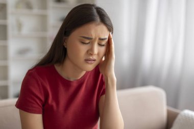 Şiddetli baş ağrısı çeken Asyalı kadın evde kendini kötü hissediyor. Genç Koreli kadın elini şakağına bastırıyor ve kaşlarını çatıyor. Oturma odasındaki koltukta oturuyor.