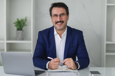 Masasında dizüstü bilgisayarı ve akıllı telefonu olan, temiz bir ofis ortamında mavi takım elbiseli, kendine güvenen beyaz bir işadamı. İş, iş.