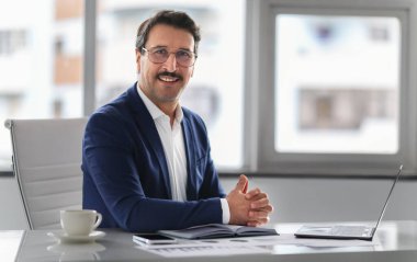 Mavi takım elbiseli, kendinden emin beyaz bir işadamı, elleri kelepçeli, kameraya gülümseyen bir dizüstü bilgisayar, bir fincan kahve ve arka plandaki belgelerle masasında oturuyor.