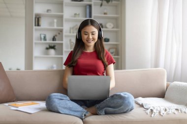 Mesafe öğrenme. Kablosuz kulaklıklı, gülümseyen Asyalı genç kadın evde dizüstü bilgisayarla ders çalışıyor, online eğitim için bilgisayar kullanan Koreli kadın oturma odasında koltukta oturuyor.