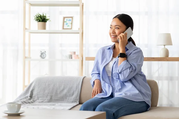 Relaxed attractive millennial chinese woman wearing comfy clothing resting on couch at home, talking on phone with friend or lover, looking at copy space. Communication concept