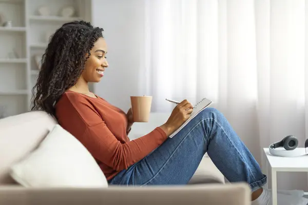 Happy young black lady drinking coffee and writing in notepad while relaxing at home, smiling african american woman enjoying cup of tea and taking notes, filling diary or making checklist, side view