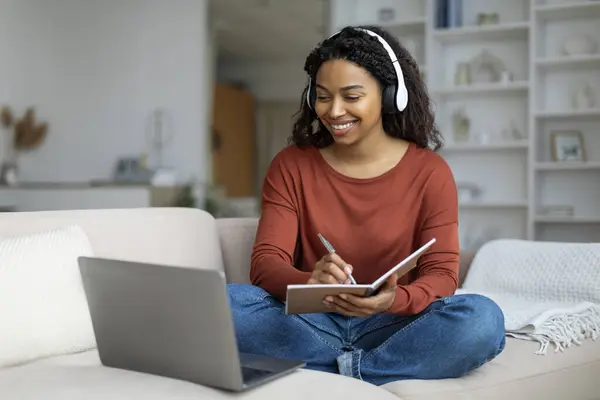 Young Black Woman In Headphones Study Online With Laptop From Home, Smiling African American Female Watching Webinar On Computer And Taking Notes To Notepad, While Sitting On Couch In Living Room