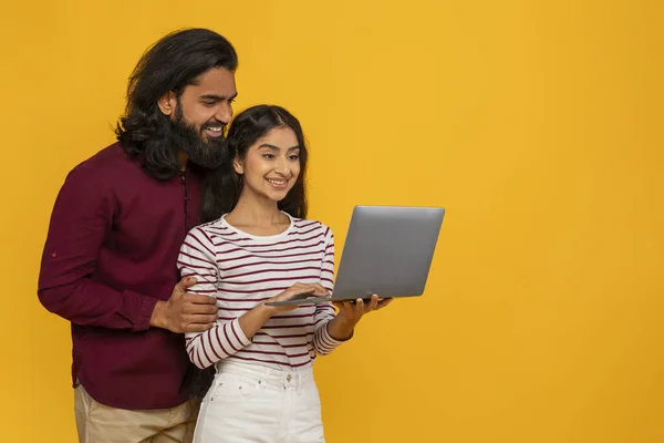 Happy millennial indian couple posing with laptop computer isolated on yellow studio background, websurfing together, copy space for advertisement, online banking, shopping. E-commerce, retail