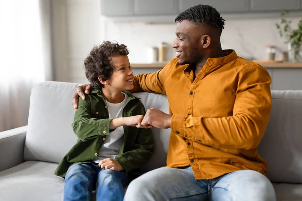 Black father and his young curly-haired son exchange joyful fist bump, sharing moment of mutual respect and understanding while sitting on the sofa