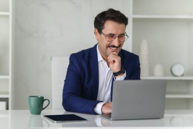 Modern bir ofis ortamında masanın üzerinde bir fincan kahve ve tabletle dizüstü bilgisayarına bakarken gülümseyen mavi takım elbiseli mutlu beyaz iş adamı. İş, iş.