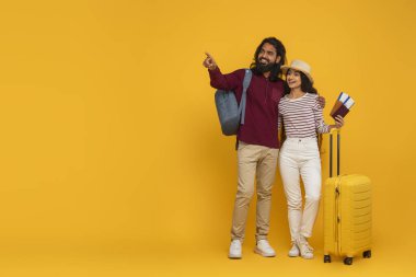Heyecanlı genç Hintli çift tatile çıkıyor, sırt çantası, çanta, pasaport ve bilet taşıyor, boş fotokopi alanını işaret ediyor, seyahat teklifini gösteriyor, sarı arka plan