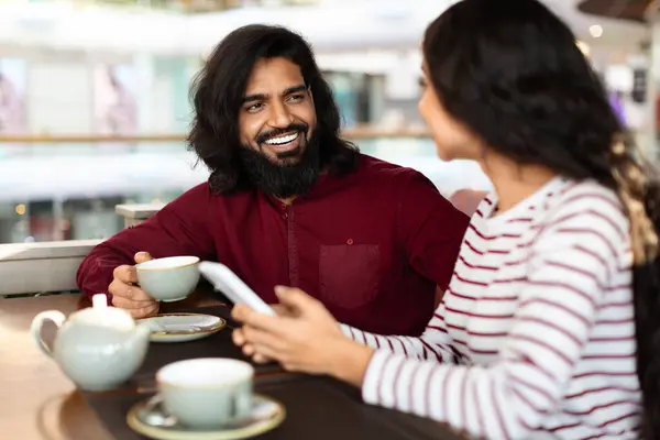 Beautiful young indian couple have romantic date at cafe, drinking tea, have conversation, using smartphone, sharing funny content, make plans. Relationships, love, affection