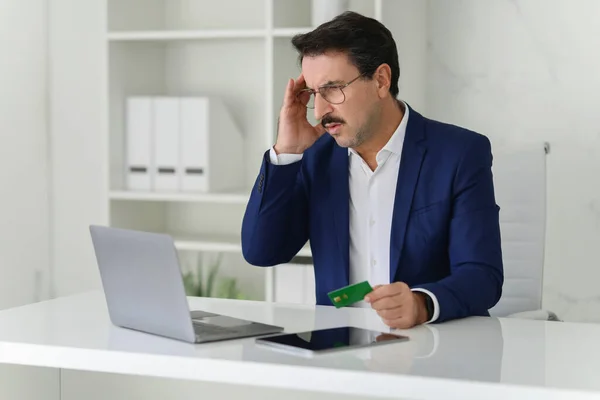 A concerned sad confused caucasian businessman in a blue suit looks stressed while holding a credit card and examining his laptop in a white modern office. . Work, business problems