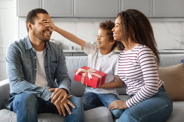 Son And Wife Greeting Young Black Man With Fathers Day, Giving Gift At Home. Male Kid Covering Dads Eyes, Surprising With Present, African American Family Sitting On Couch In Living Room, Closeup