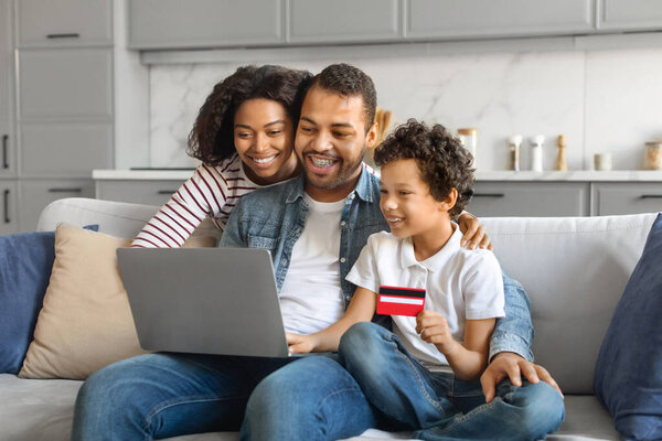 Online Shopping Concept. Happy Black Family With Little Son Using Laptop And Credit Card At Home, Buying Goods In Internet, African American Parents And Their Child Sitting With Computer On Couch