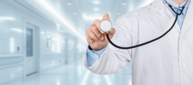 Beyaz önlüklü ve mavi gömlekli bir tıp uzmanı modern bir hastane koridorunda kameraya steteskop uzatarak klinikte ve panoramada bakım sağlamaya hazır olduğunu gösteriyor.