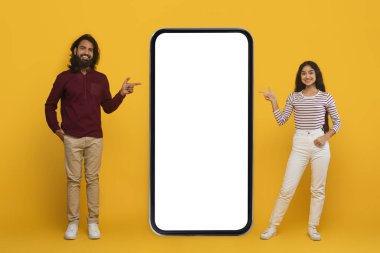 Harika bir online teklif, anlaşma, güzel mobil uygulama. Gülümseyen genç Hintli çift, sarı arka planda izole edilmiş beyaz ekranlı akıllı telefonu işaret ediyor.