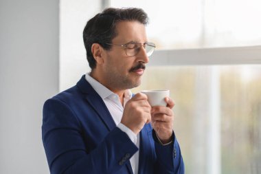 Mavi takım elbiseli, gözlüklü, olgun beyaz bir işadamı taze bir fincan kahvenin tadını çıkarıyor. Parlak bir ofisin penceresinde huzurlu bir anın tadını çıkarıyor.
