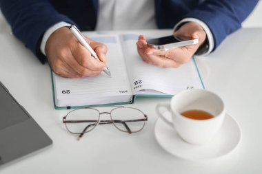 Kafkasyalı bir iş adamının elleri gösteriliyor, biri açık bir planlayıcıyla, diğeri ise akıllı bir telefonla, masasında gözlük ve bir fincan çay ile, yoğun bir iş günü olduğunu gösteriyor.