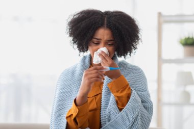 Kafası karışık, kıvırcık saçlı, termometreyle ateşini ölçen, kendini kötü hisseden, örgü örülmüş mavi bir battaniyeye sarılı Afro-Amerikalı kadın.