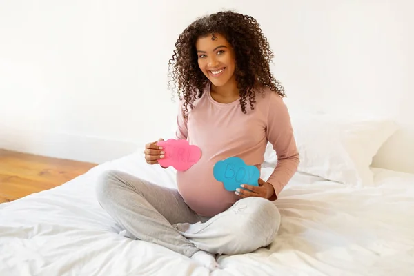 GirlとBoyを読んでピンクとブルーのサインでベッドに座っていることを期待して陽気な 幸せな若いアフリカ系アメリカ人妊婦は 性別のために彼女の興奮を分かち合います — ストック写真