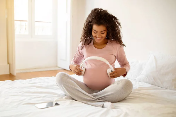 Black Pregnant Woman Holding Headphones Near Belly, Playing Melody To Baby In Womb, Happy Lady Enjoying Listening Music During Pregnancy, Sitting On Bed At Home, Closeup Shot With Free Space