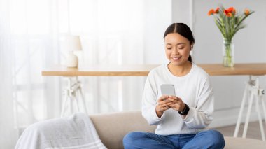 Mobil uygulamayı eğlendiriyorum. Neşeli Asyalı kadın evdeki rahat oturma odasındaki kanepede dinleniyor, akıllı telefon ve kablosuz kulaklık kullanıyor, favori podcast izliyor, müzik dinliyor, yer kopyalıyor.