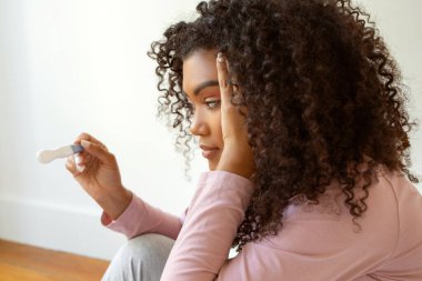 Endişeli genç siyahi kadın yerde oturuyor ve hamilelik testi yapıyor. Afro-Amerikan kadın sonuçtan endişeli ve dalgın görünüyor. Karmaşık duygular hissediyor, yakın çekim yapıyor.