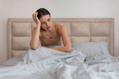 Sorunlu genç bir kadın yatakta oturuyor, eli başının üstünde, endişeli görünüyor, muhtemelen baş ağrısı ya da bunaltıcı düşüncelerle uğraşıyor, kopyalama alanı